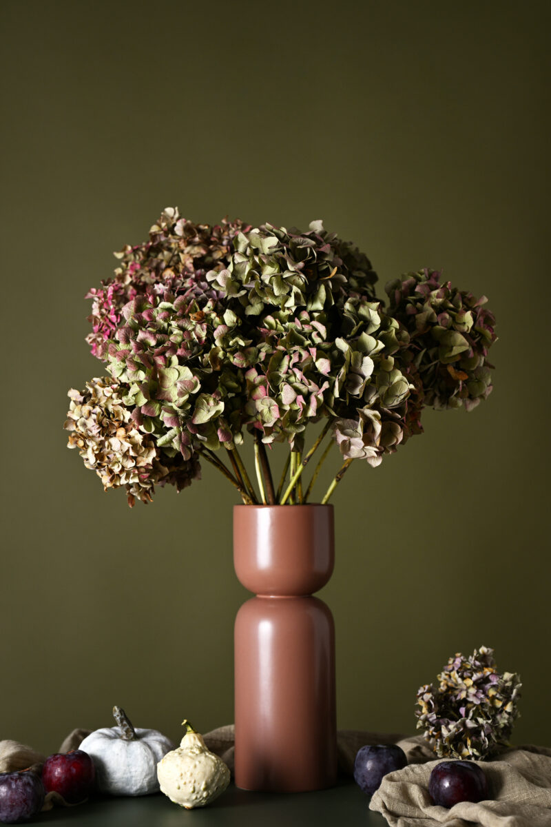 Dried hydrangea flowers arranged in a tall vase with decorative pumpkins and fall accents.